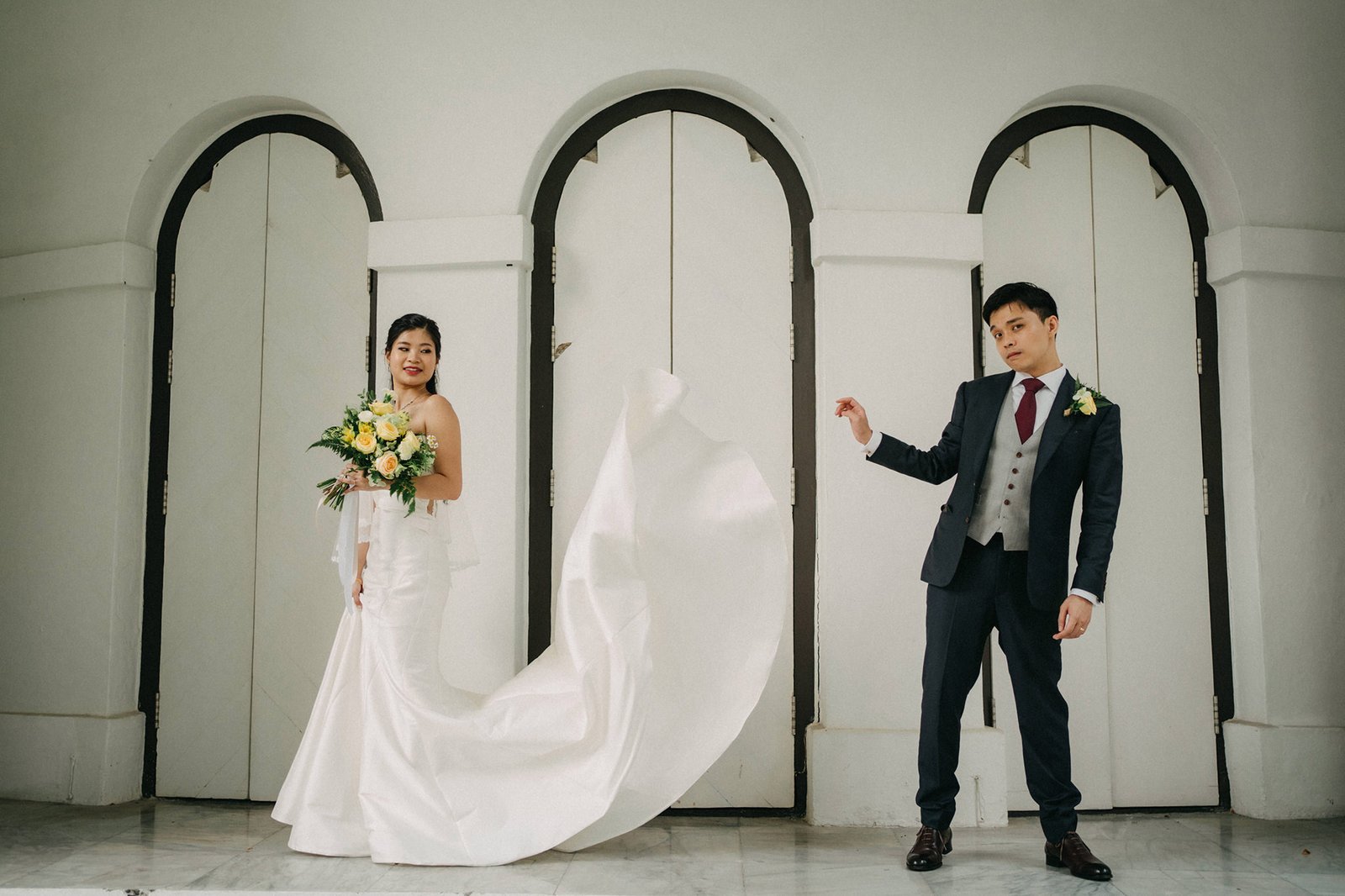 A smiling bride in a white strapless gown holds a bouquet of yellow and white flowers, while a groom in a dark suit playfully lifts her veil in front of arched doorways.