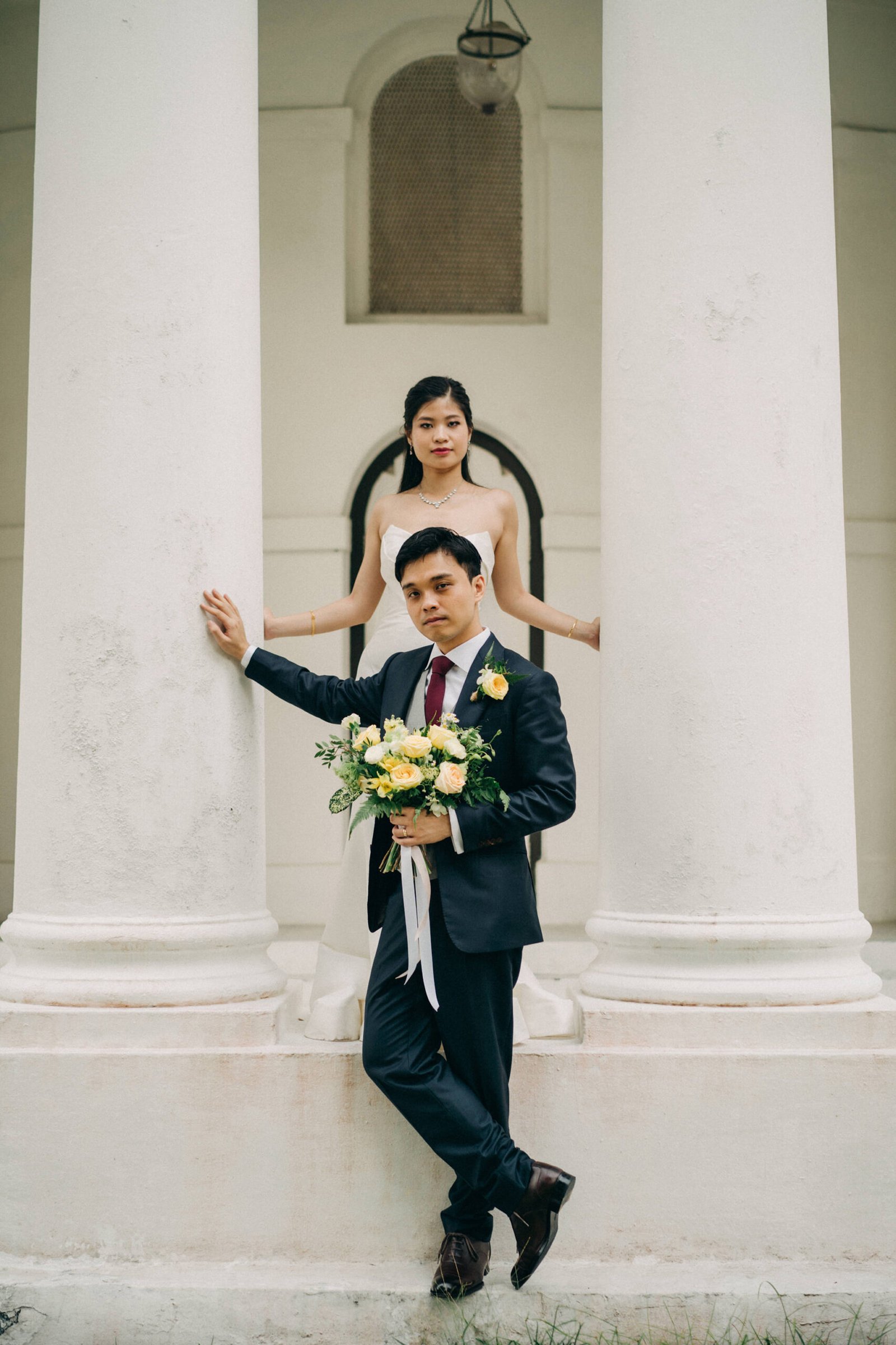 A bride and groom pose between large white columns at an outdoor wedding venue. The groom in a dark suit holds a bouquet of yellow and white flowers.