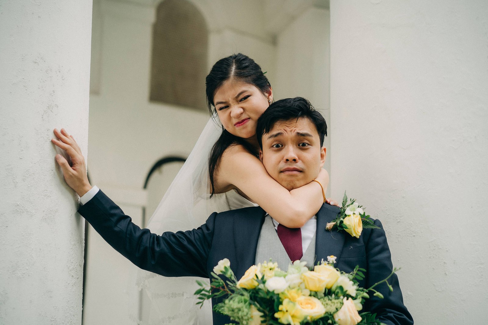 A bride playfully chokes a groom in a wedding photo, both with exaggerated expressions. The groom holds a bouquet of yellow and white flowers.
