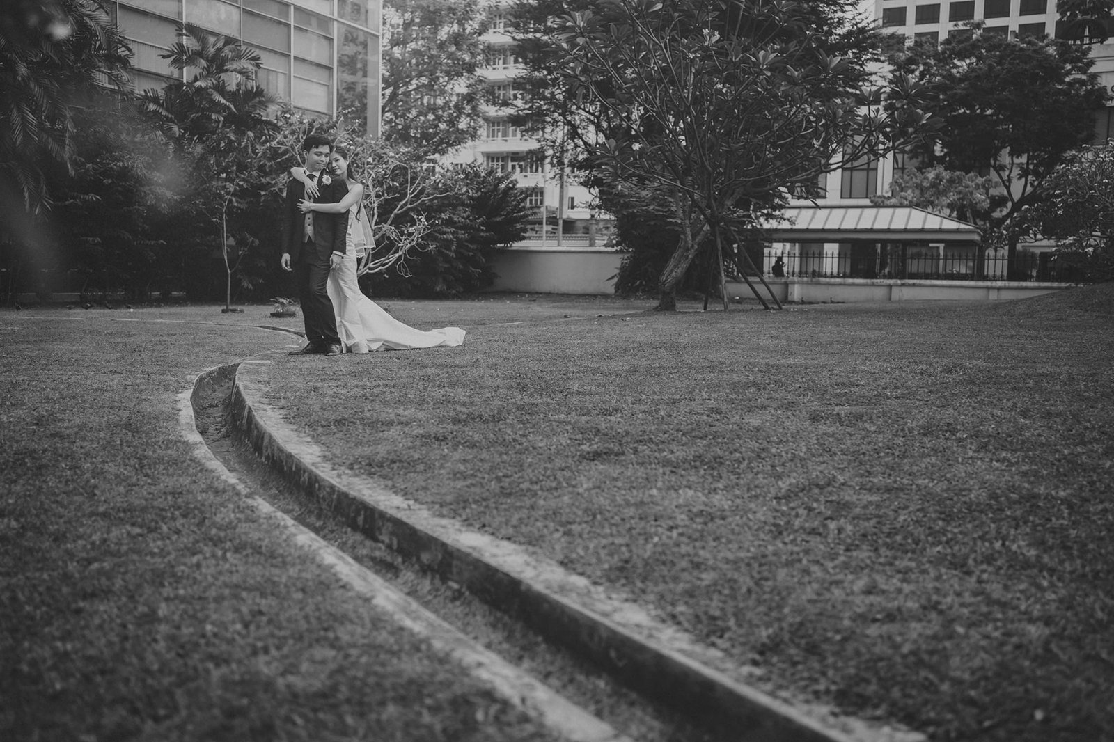 Black and white photo of a wedding couple embracing in a park with lush green grass and trees.
