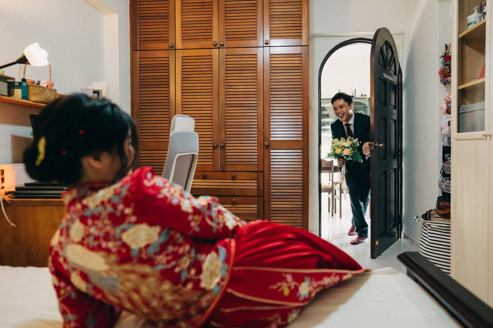 A smiling groom in a suit holds a bouquet of flowers as he enters a bedroom where his bride, in a red traditional Chinese wedding dress, turns to greet him.
