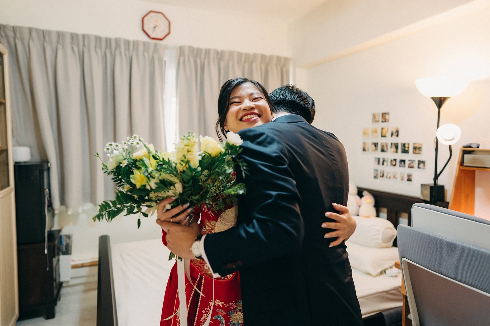 A joyful bride in a red traditional dress embraces her groom in a suit, holding a beautiful bouquet of yellow and white flowers. The scene appears to be a wedding morning in a bedroom.