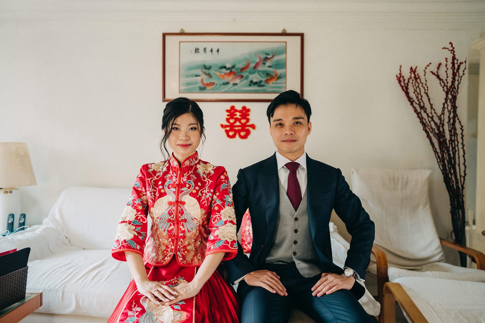 A smiling Asian bride in a red traditional Chinese wedding dress and a groom in a dark suit and vest sit side-by-side on a couch, with a "囍" (double happiness) symbol and a traditional painting on the wall behind them.