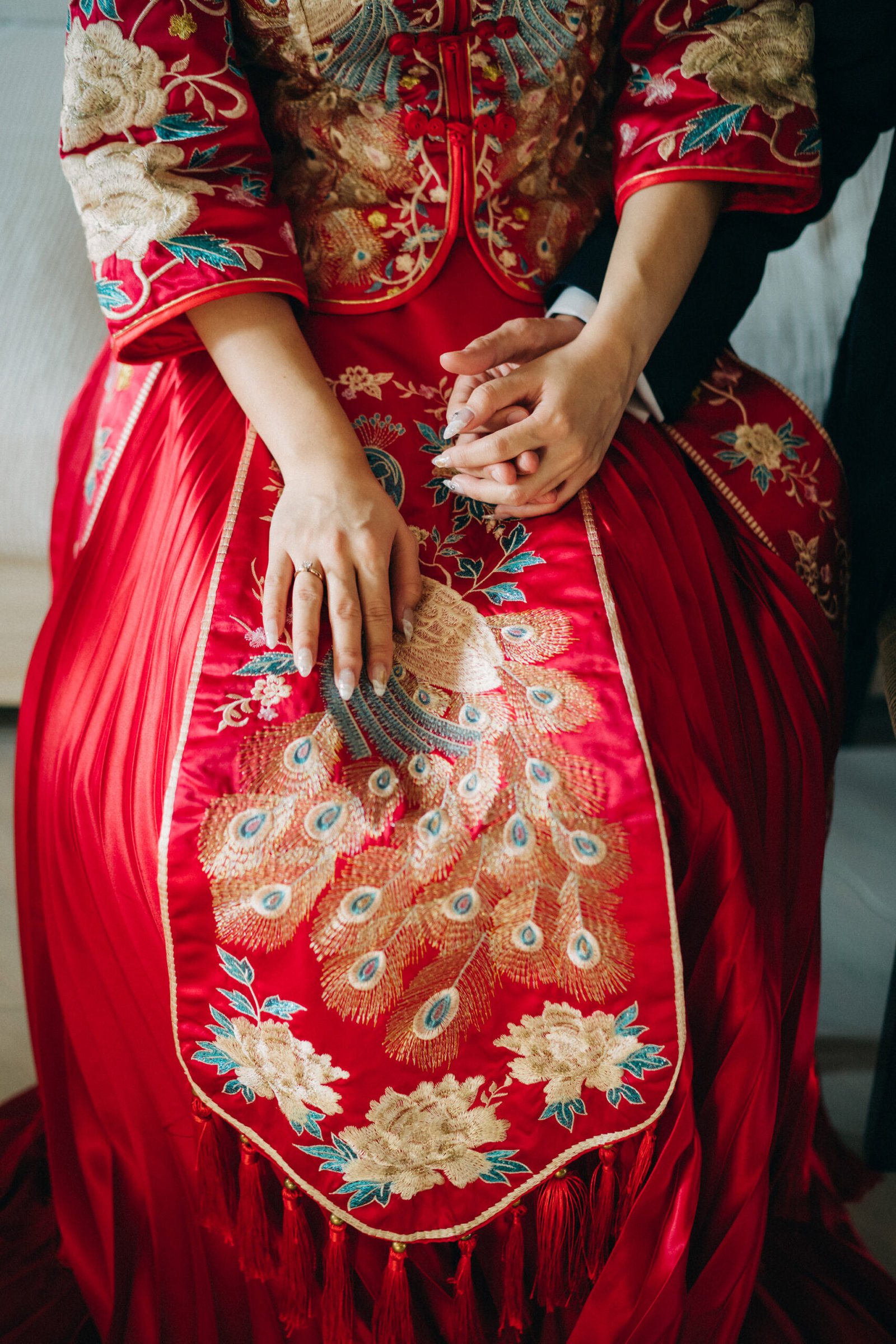 Close-up of a bride in a traditional red Chinese wedding dress adorned with intricate gold embroidery of peacocks and floral patterns. Her hands, wearing a wedding ring, are clasped with her partner's hands.