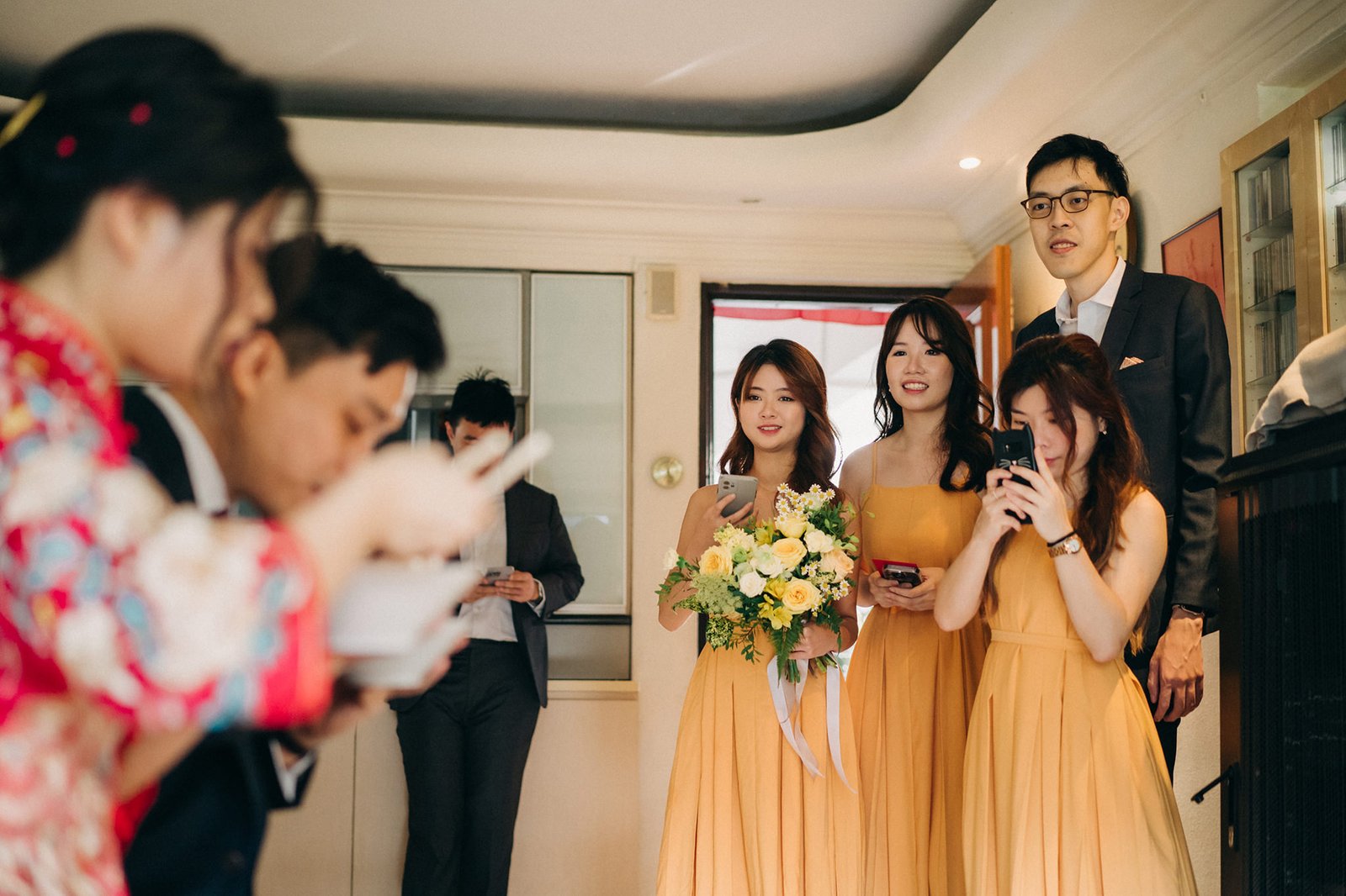 A wedding party in a bright room, with bridesmaids in yellow dresses holding phones and a bouquet, while guests in the foreground are blurred and looking at their phones.