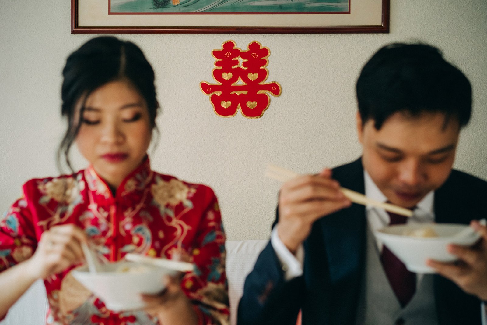 A Chinese bride in a red traditional dress and a groom in a suit eat from bowls with chopsticks, with a red double happiness symbol on the wall behind them.