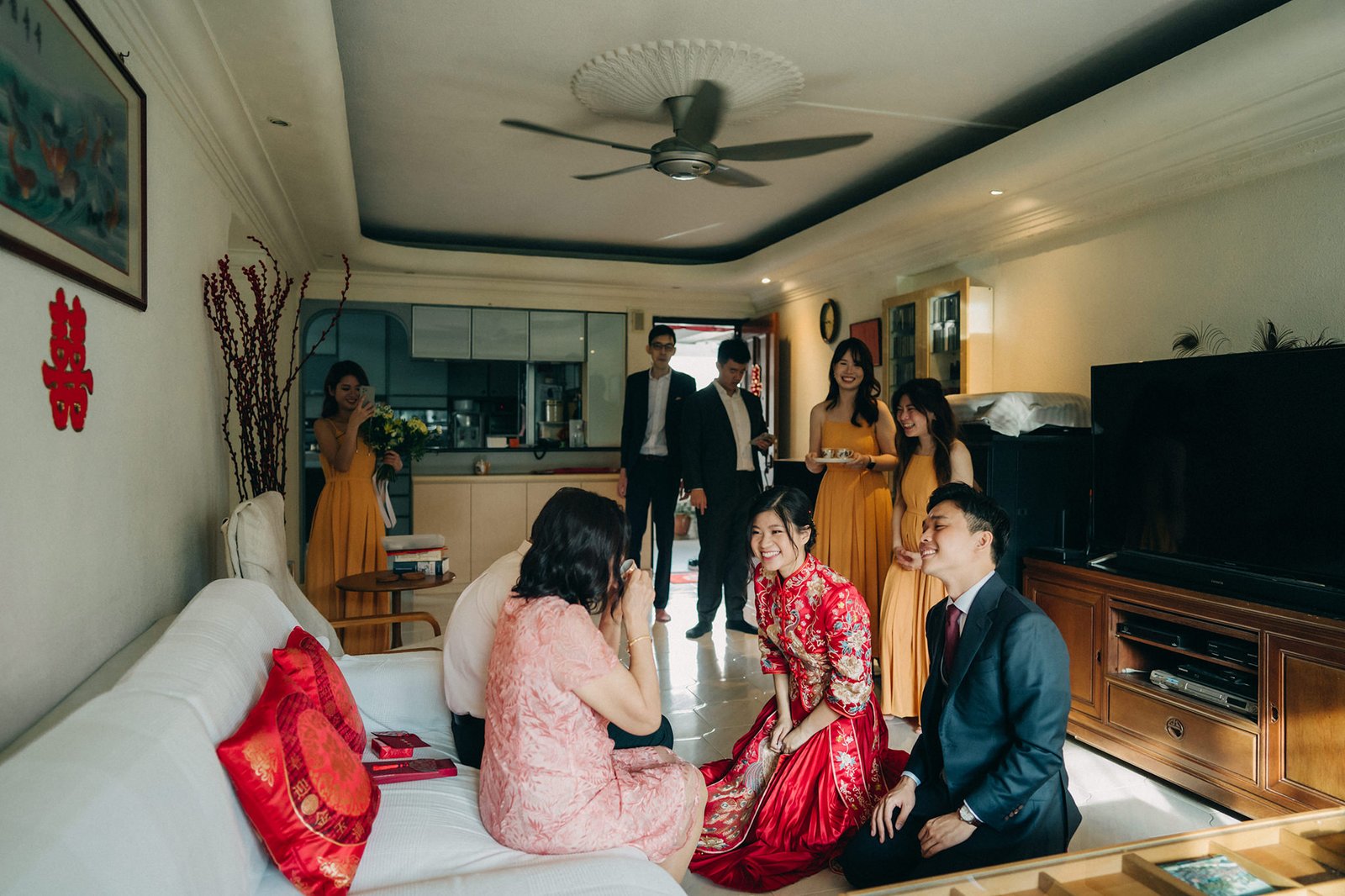 A joyous Chinese wedding ceremony unfolds indoors, with a bride in a stunning red cheongsam and groom in a dark suit kneeling on the floor, surrounded by family and bridesmaids in yellow dresses.