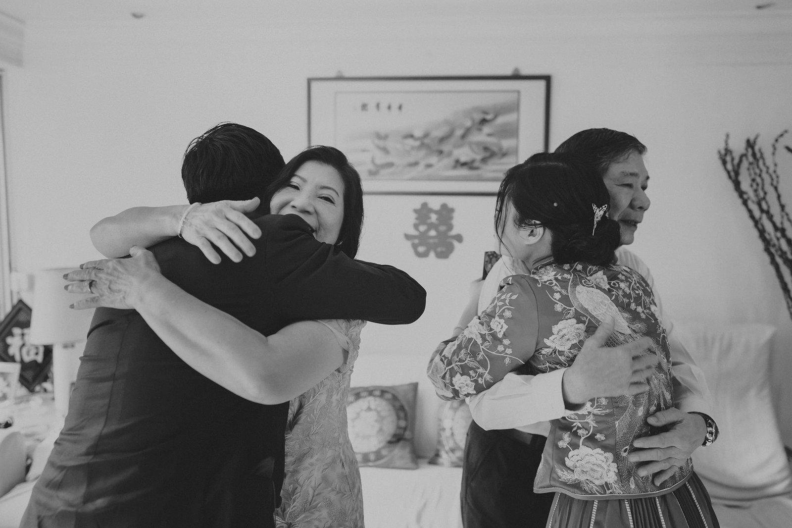 Black and white photo of two couples embracing, a bride in a traditional Chinese dress hugging her parents, with a wedding painting in the background.