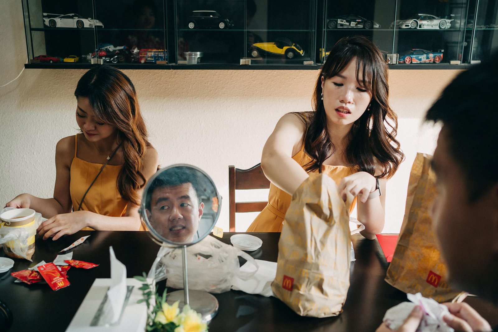 Two young women in yellow dresses sit at a table, with one woman holding a McDonald's bag. A mirror reflects a man's face in the foreground. Shelves with toy cars are visible in the background.