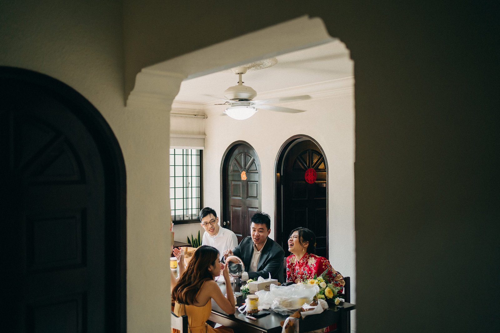 A joyful wedding preparation scene: a bride in a vibrant red traditional Chinese dress laughs while a groom and two groomsmen gather around a table, with makeup and floral arrangements visible. This is a glimpse into a special wedding day.