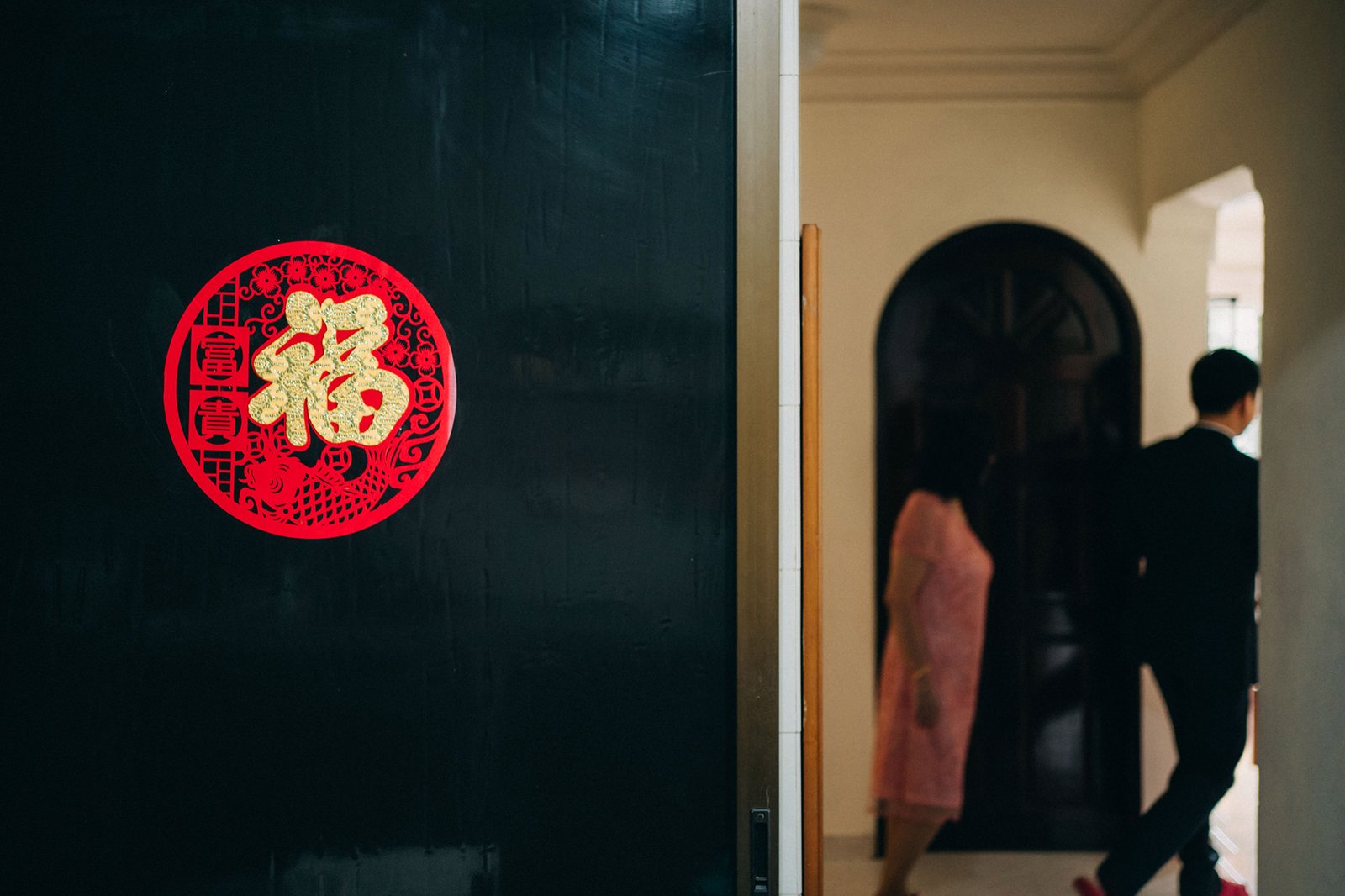 A red and gold Chinese "Fu" character paper cut decoration is affixed to a dark, textured wall, with a blurred couple in formal attire walking through an arched doorway in the background.