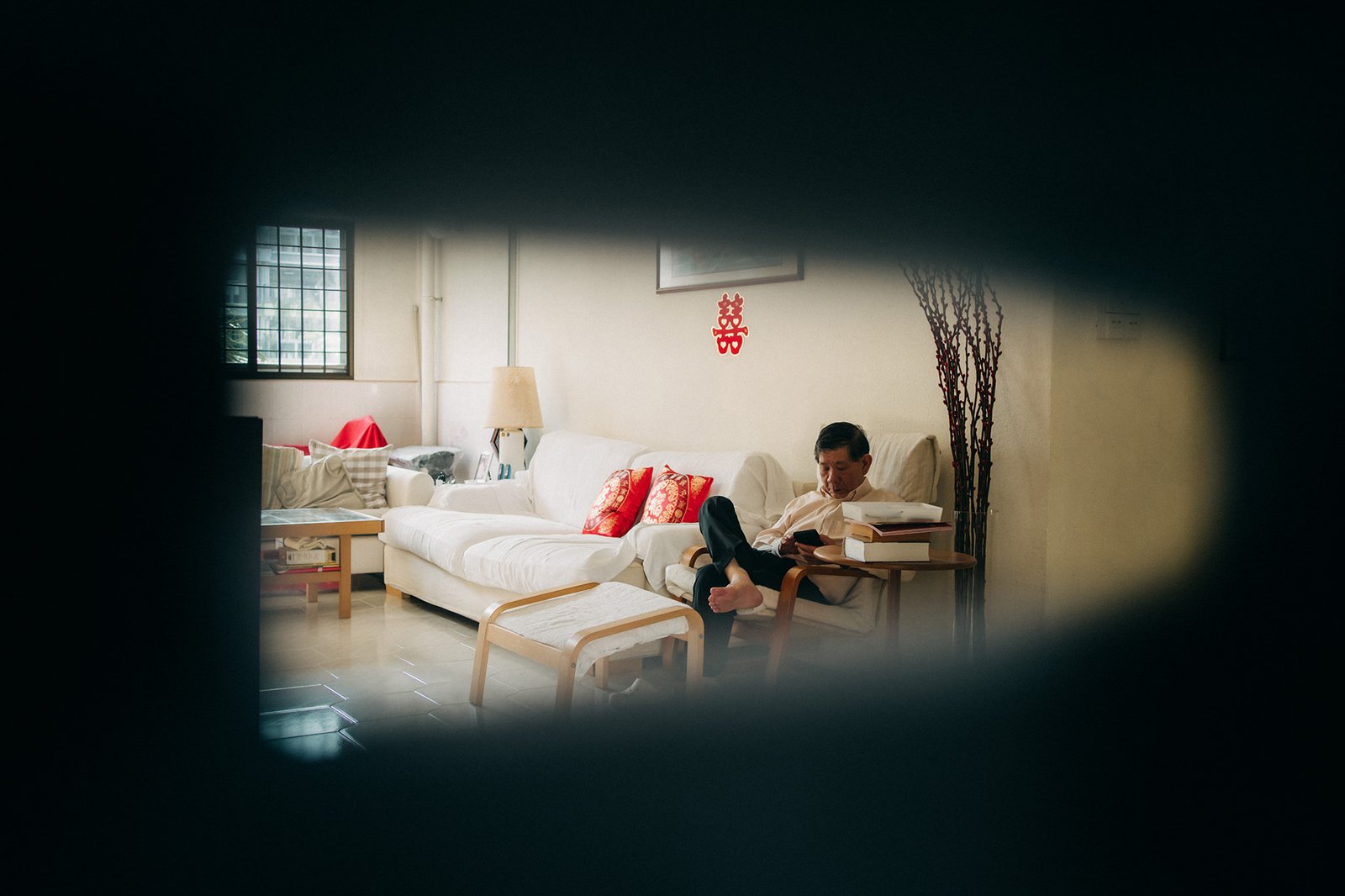 An older Asian man relaxes on a white sofa in a living room, scrolling on his phone. A red double happiness symbol adorns the wall behind him.