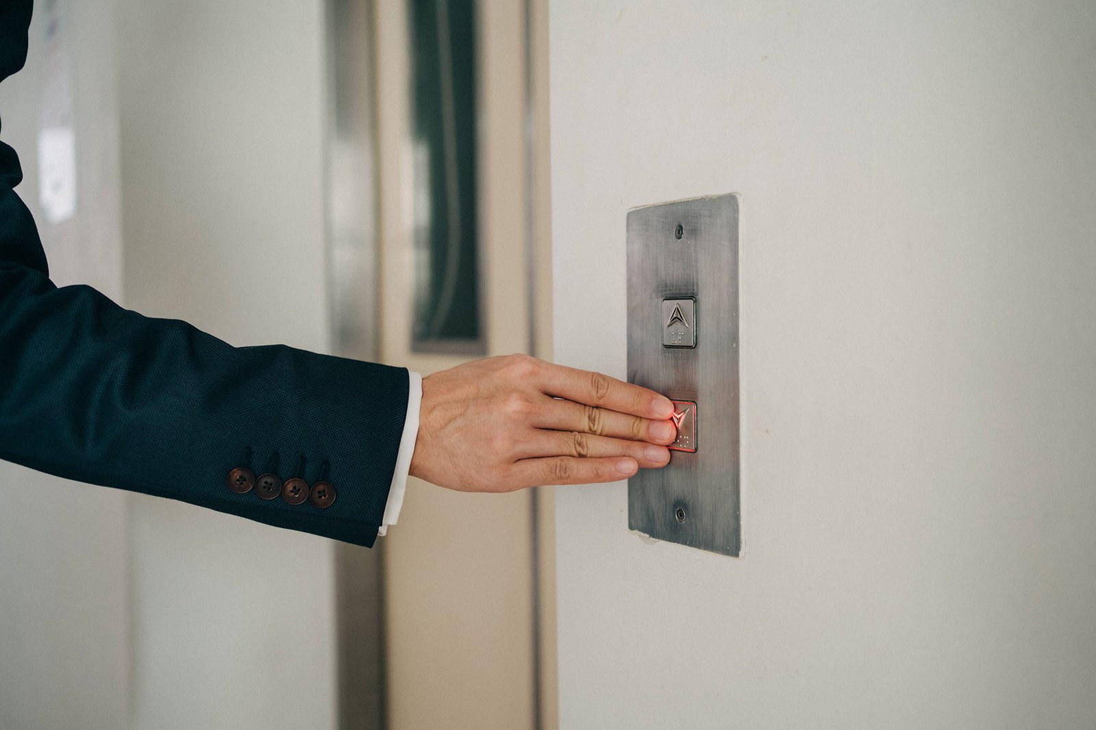 A person in a dark suit presses the illuminated red elevator call button on a textured wall.