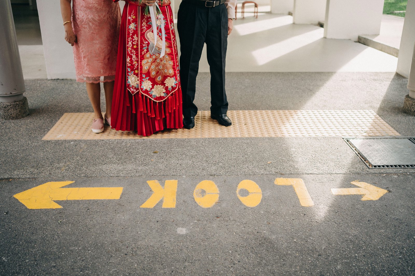Three people stand on an outdoor walkway with a yellow "LOOK" sign and arrows painted on the ground. The person in the middle wears a traditional red and gold Chinese wedding dress.