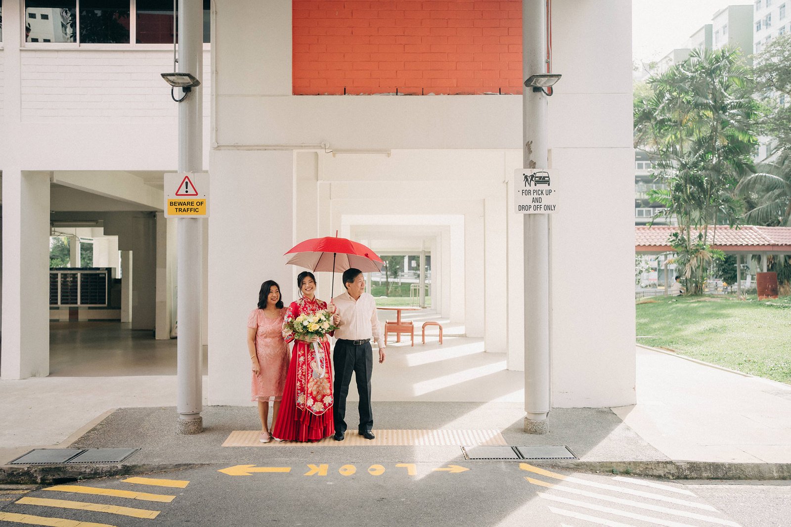 A family of three, a bride in a red traditional Chinese dress, her parents, pose under a red umbrella outside a modern building with white walls and orange accents. "BEWARE OF TRAFFIC" sign visible.