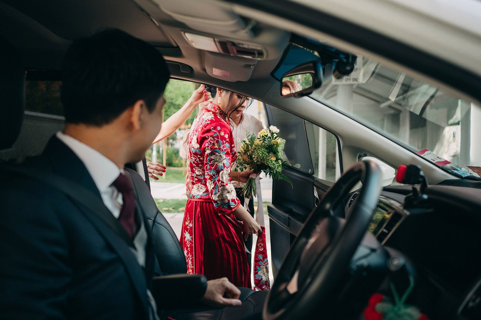 A bride in a vibrant red traditional Chinese wedding dress, holding a bouquet, is helped into a car by a bridesmaid. A man in a suit is visible in the foreground.