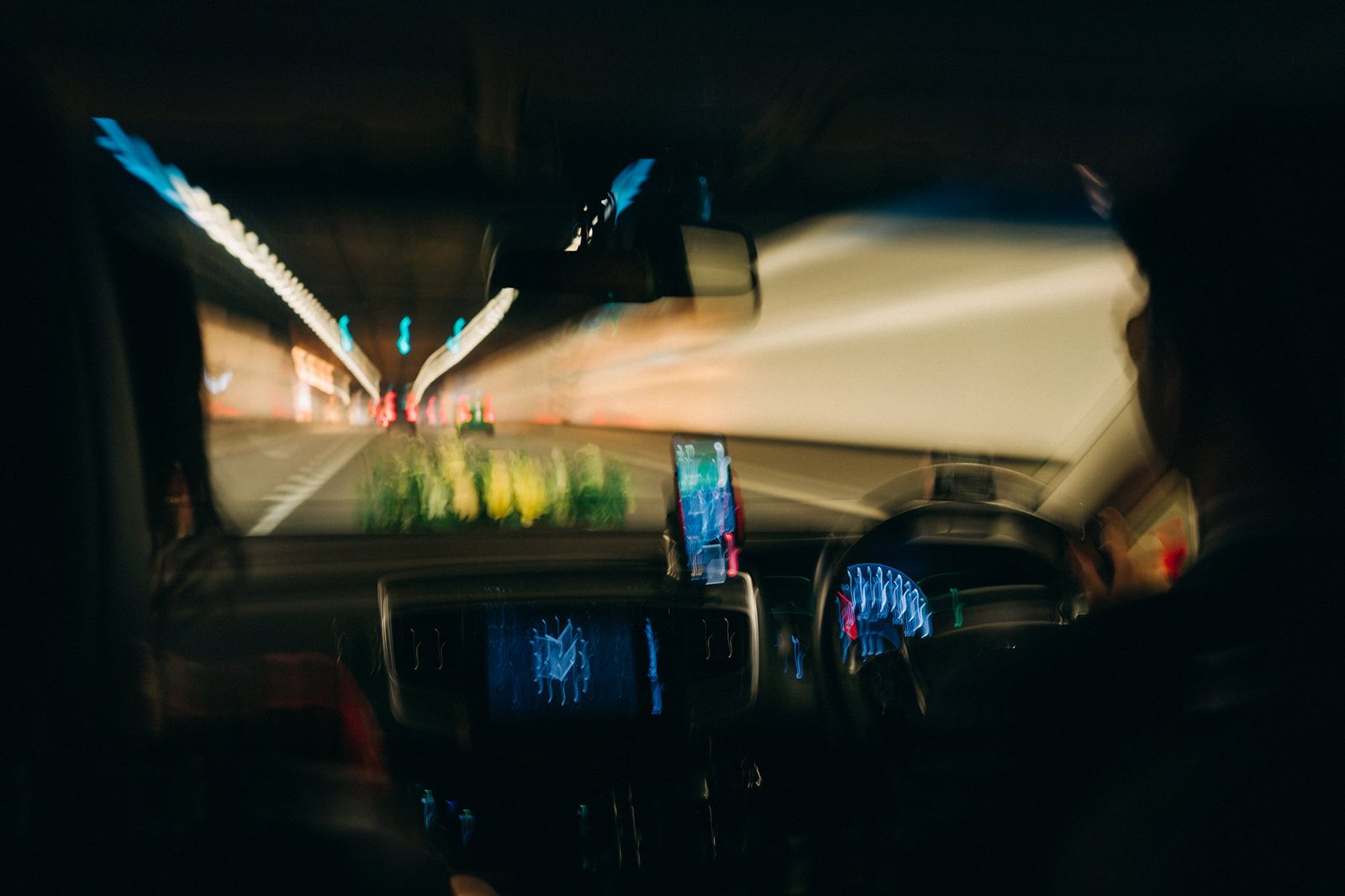 Motion blur captures a car's interior at night, with illuminated dashboard gauges, a phone mount, and the streaking lights of a tunnel or highway visible through the windshield.