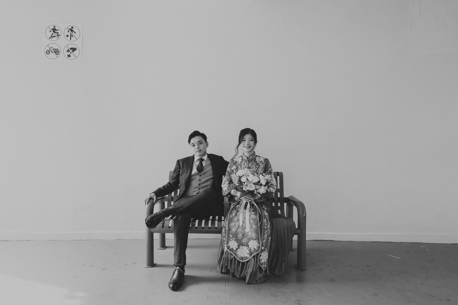A bride in a traditional Chinese wedding dress and groom in a suit sit on a bench, holding a bouquet. The black and white photo captures a formal portrait with a minimalist background.