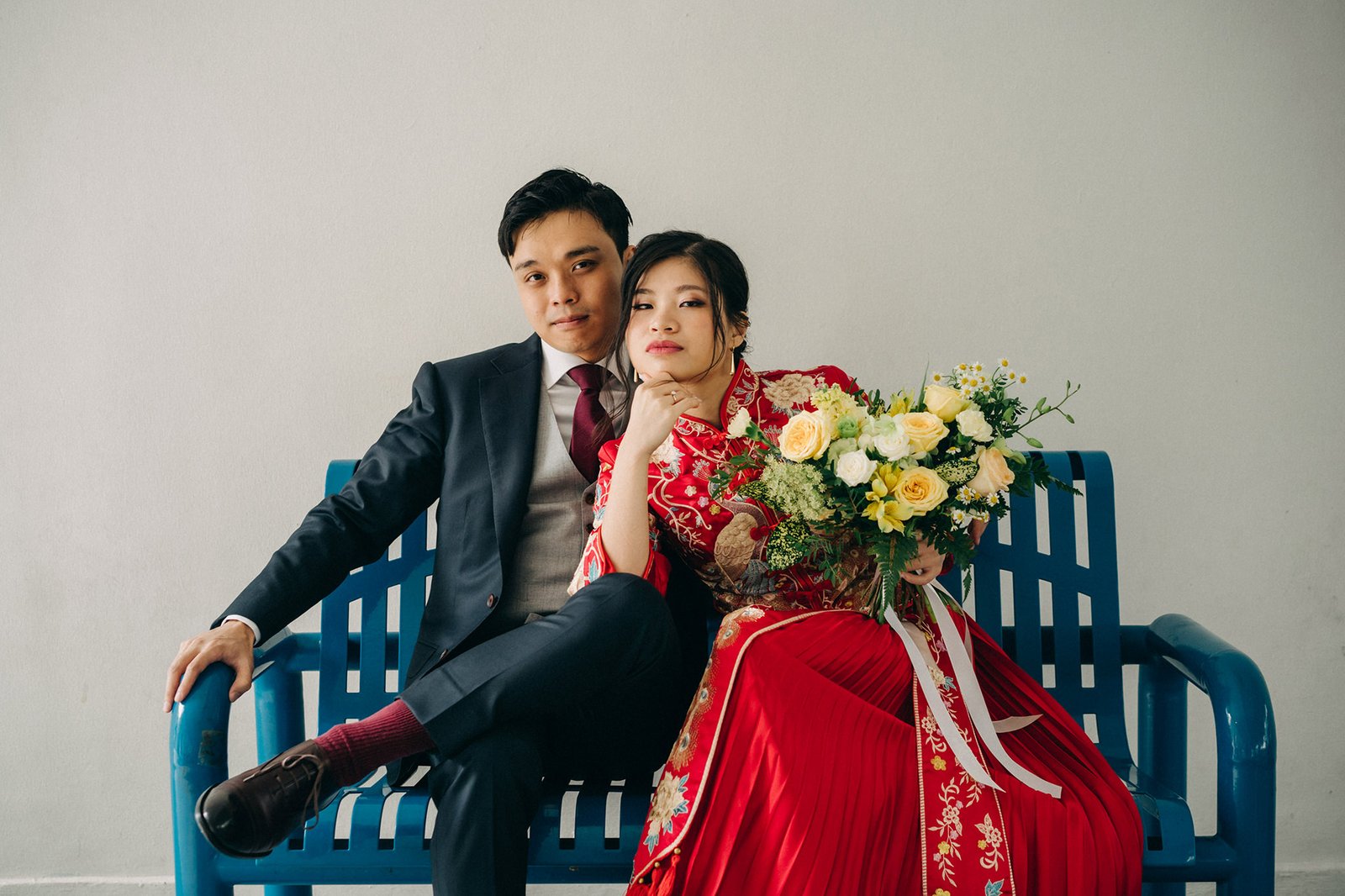A newlywed Asian couple in traditional red wedding attire sits on a blue bench, holding a vibrant bouquet of yellow and white flowers.