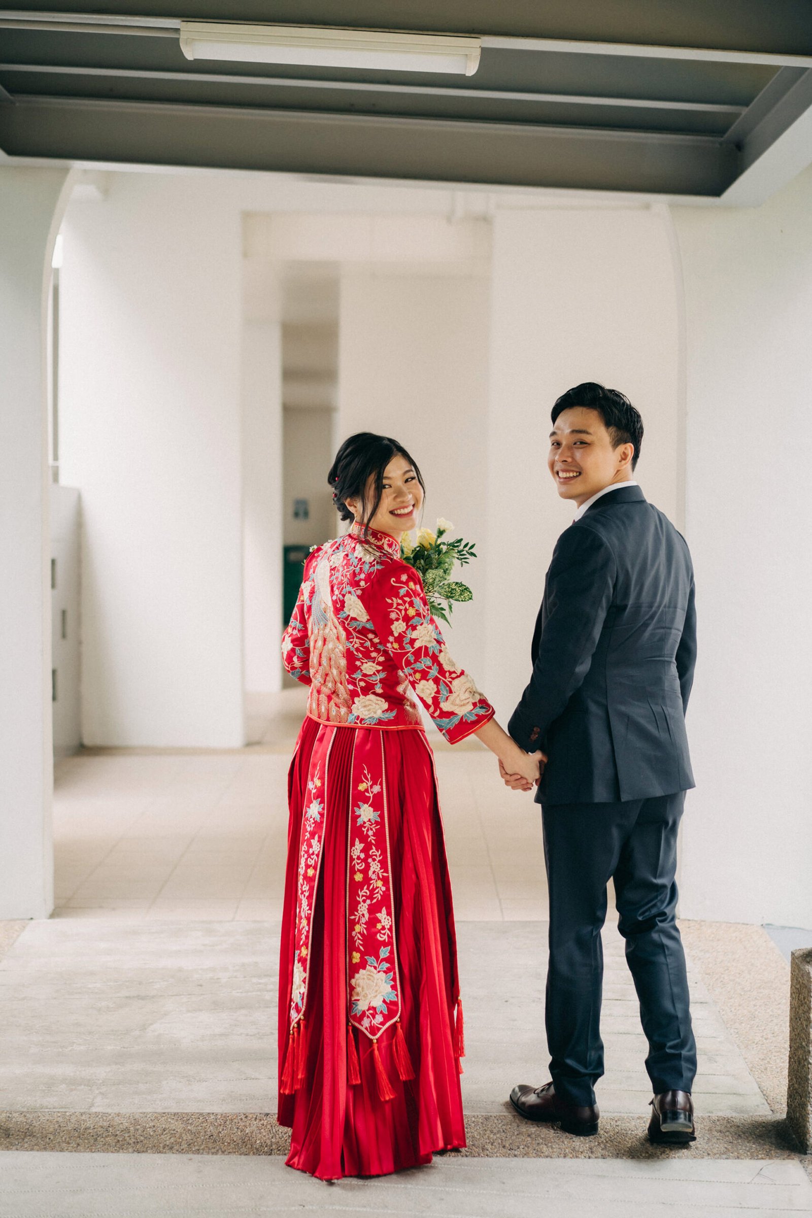 A smiling Asian couple in wedding attire, the bride in a red qipao and the groom in a dark suit, hold hands and look back over their shoulders while walking down an arched corridor.