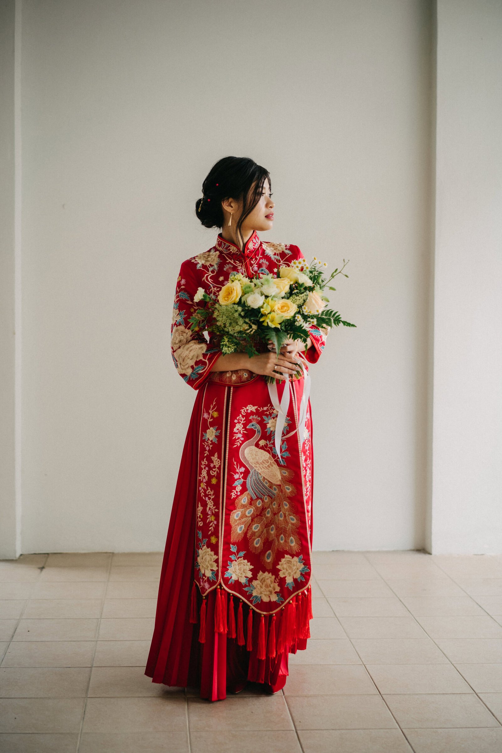 A stunning Asian bride in a traditional red Qipao, adorned with intricate gold embroidery and a peacock motif, holds a bouquet of yellow and white roses. She stands against a plain white wall, her hair styled in an elegant updo.