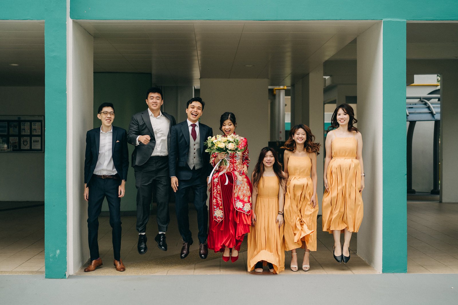 A wedding party of seven people, including a bride in a red and gold dress and a groom in a suit, joyfully jump in unison in a modern outdoor hallway.