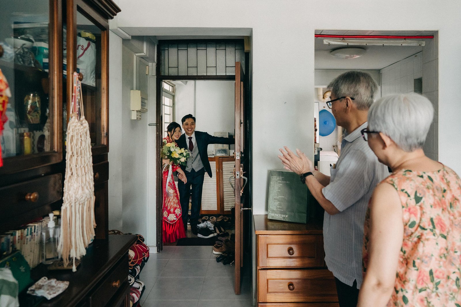 A smiling groom in a dark suit and red-trimmed traditional attire, holding a bouquet, is welcomed by an elderly couple at a doorway. The scene captures a joyous wedding moment with a warm, welcoming atmosphere.