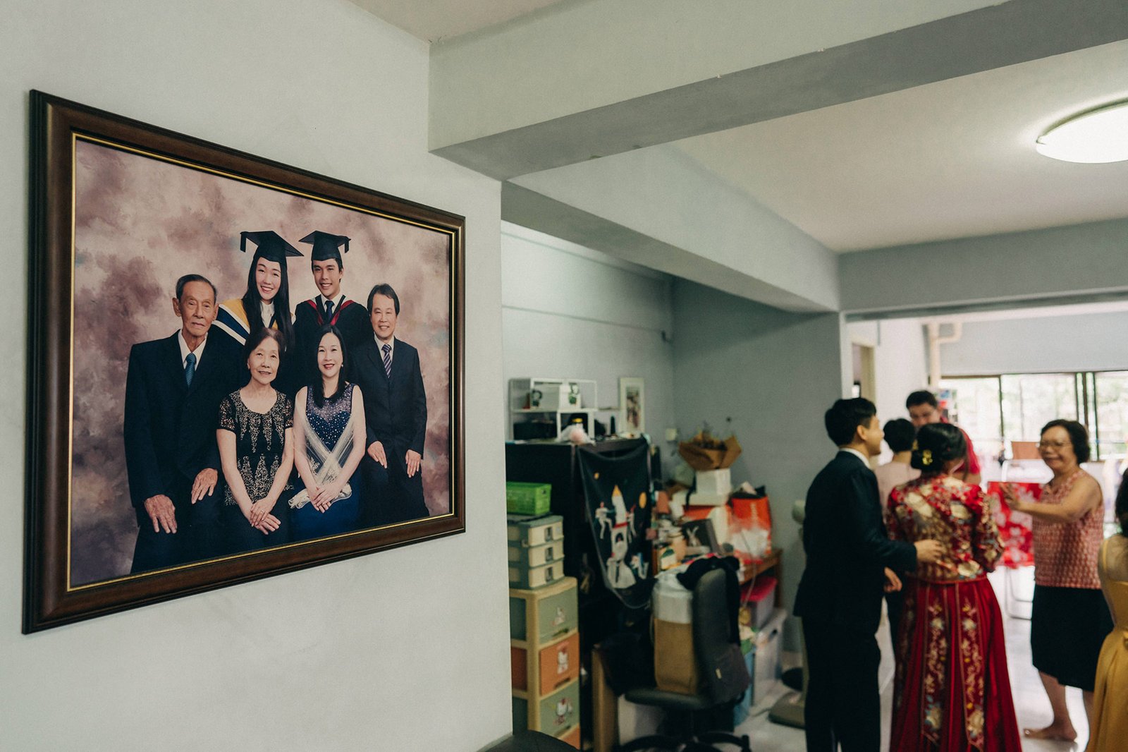 A formal family portrait in graduation caps hangs on a wall, featuring eight individuals dressed in elegant attire. In the background, a wedding ceremony is underway with guests in festive clothing.