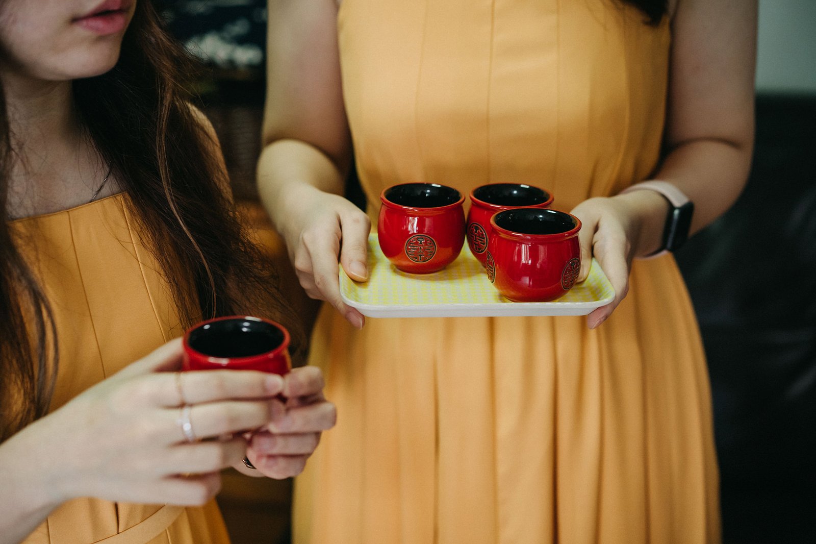 Two women in yellow dresses hold small red tea cups, one on a yellow tray with three cups, the other holding one cup. The cups have a gold Chinese symbol.
