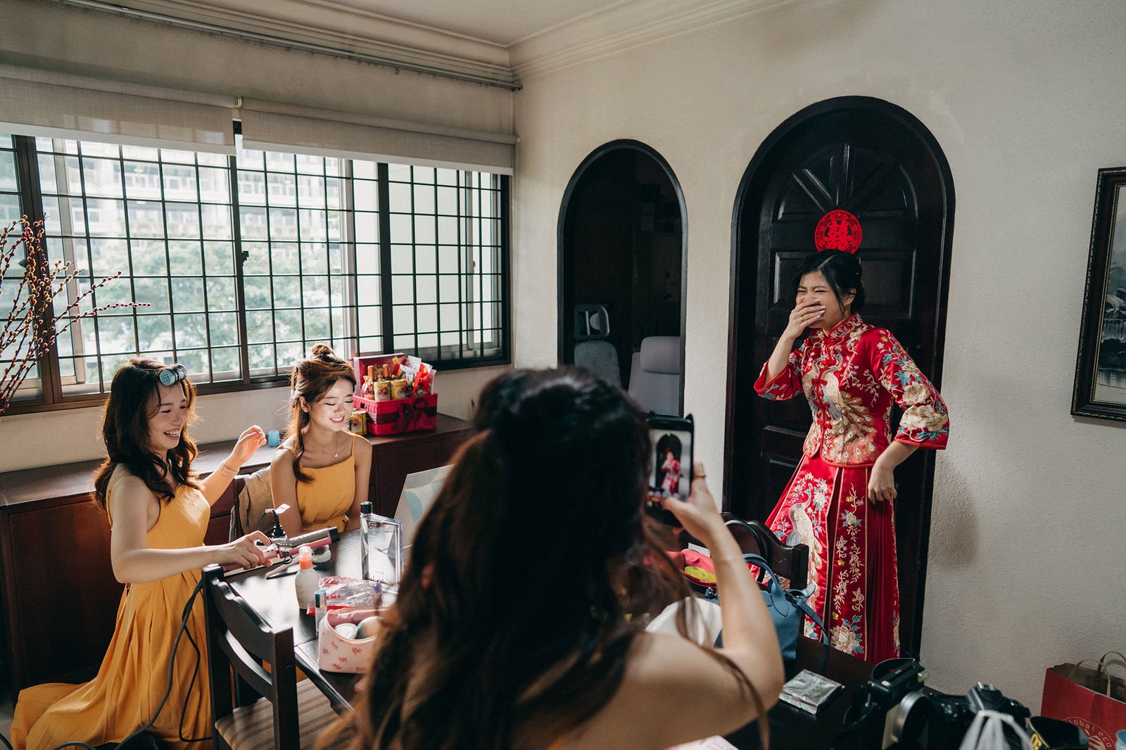 Bride in a red traditional Chinese wedding dress laughs with bridesmaids in yellow dresses, getting ready for a Chinese wedding.