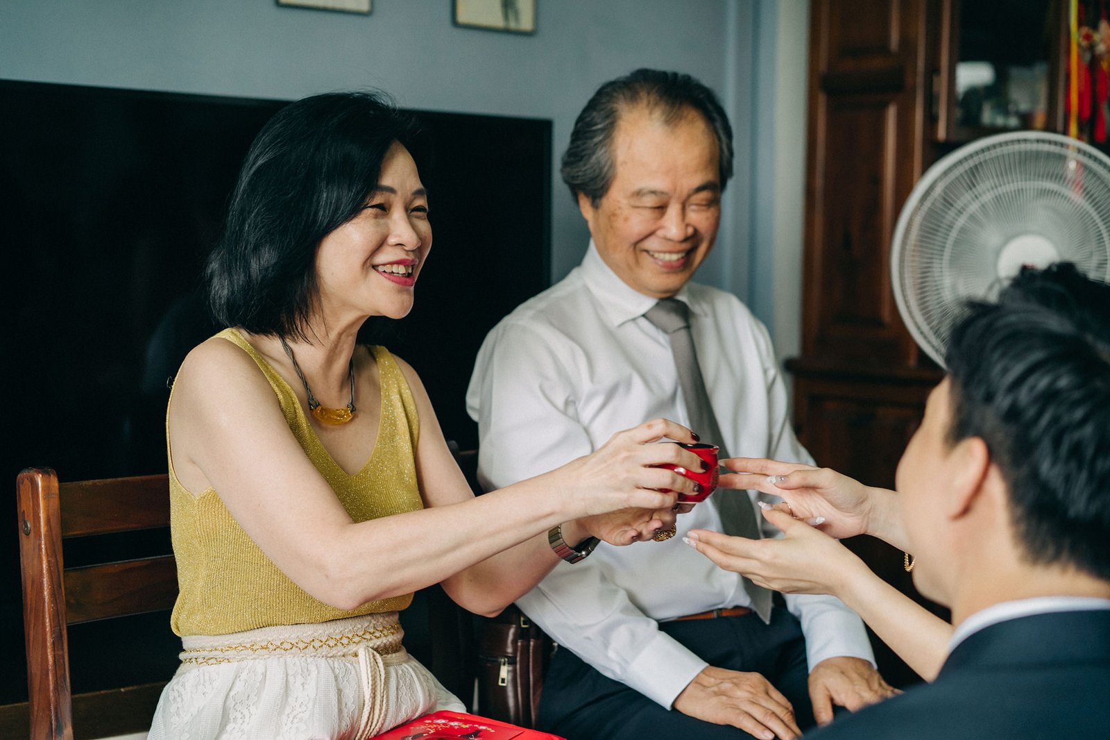 A smiling woman in a gold top offers a red teacup to a young man during a traditional tea ceremony, with an older man in a white shirt smiling in the background.