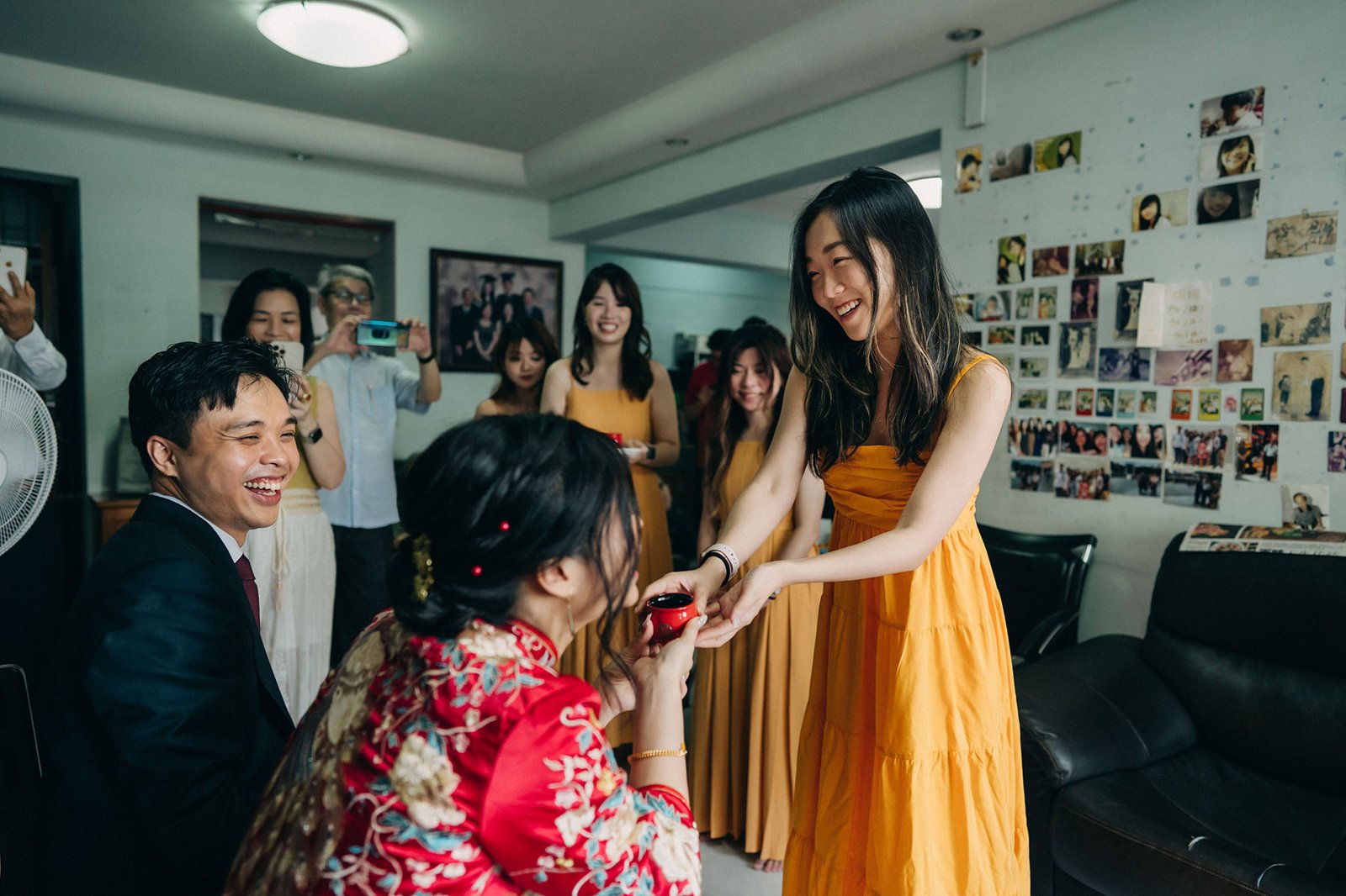 A bride in a red traditional Chinese wedding dress kneels to serve tea to a smiling woman in an orange dress during a tea ceremony, with family members watching and taking photos in the background.