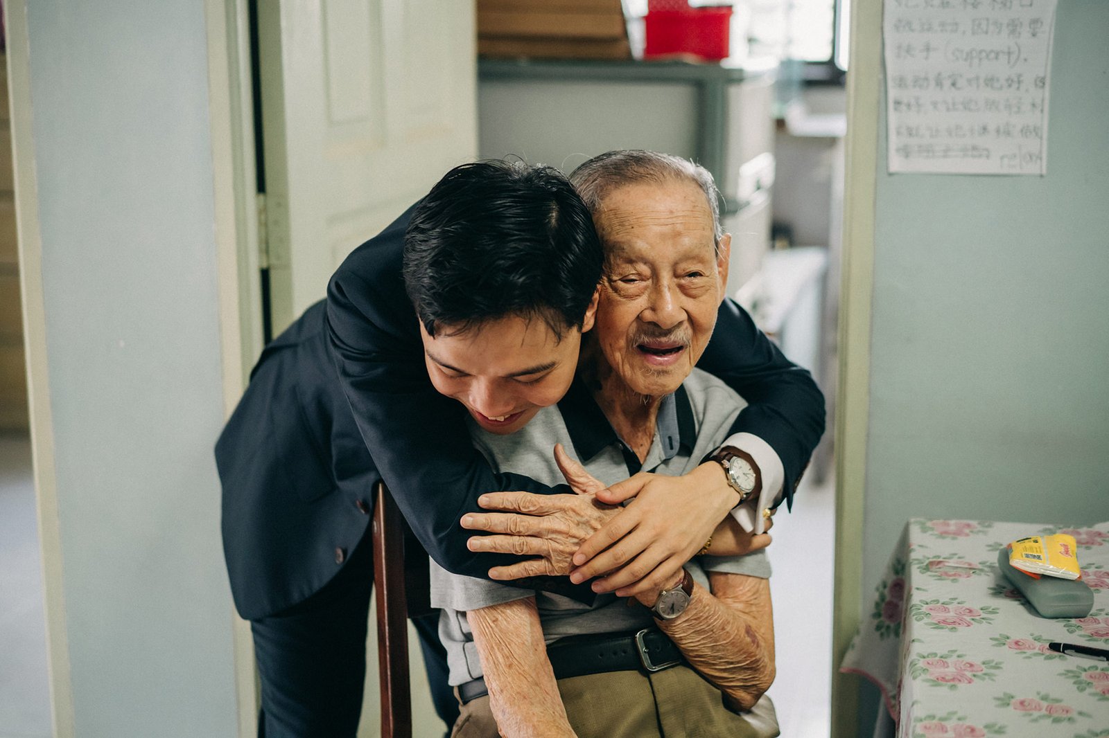 A young man in a suit hugs an elderly man sitting in a chair, with a warm embrace. The scene conveys intergenerational love and family connection.