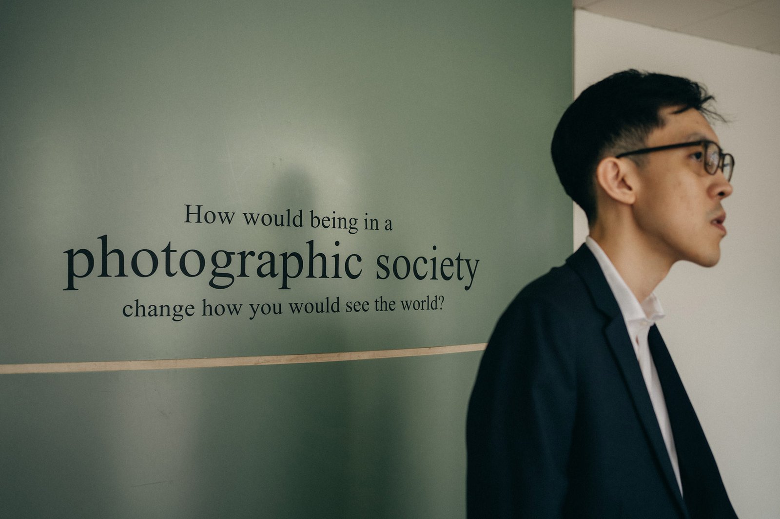 A man in a suit and glasses stands in front of a green wall with the text "How would being in a photographic society change how you would see the world?