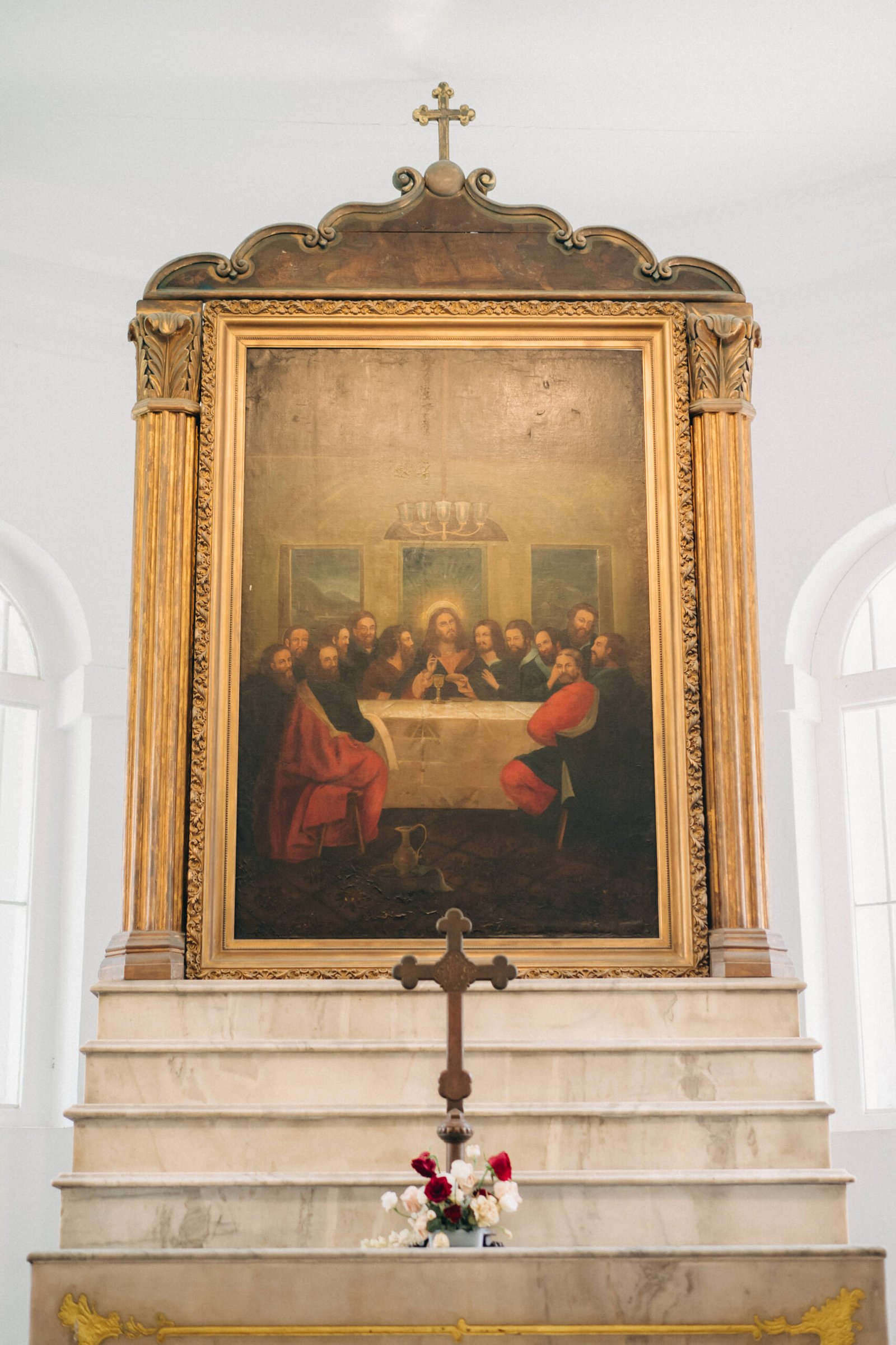 A religious painting of the Last Supper hangs in a church above a marble altar, with a wooden cross and flowers in the foreground.