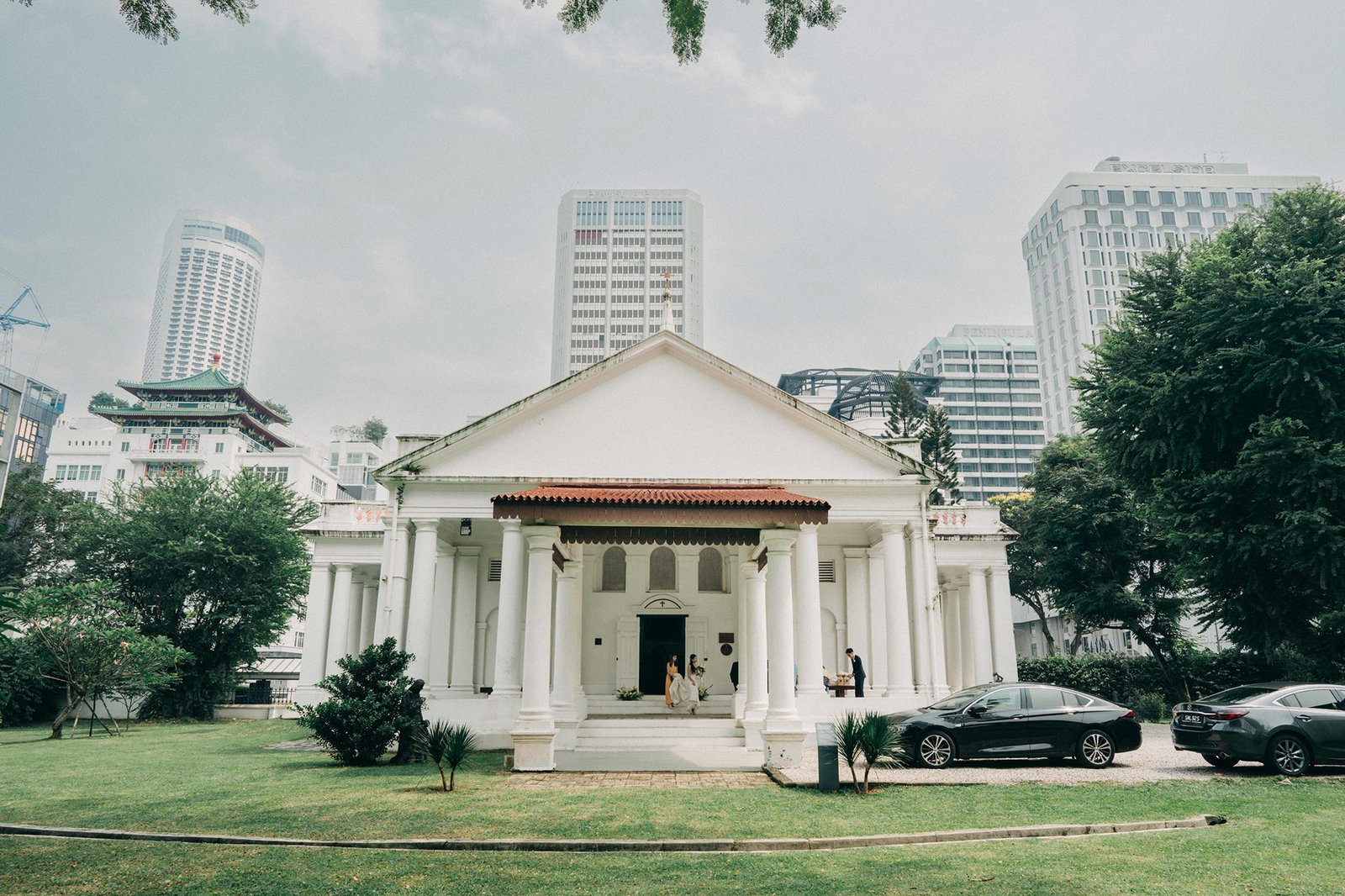 Armenian Church Singapore building with columns and a red-tiled roof stands on a green lawn, with modern skyscrapers in the background and cars parked in front.