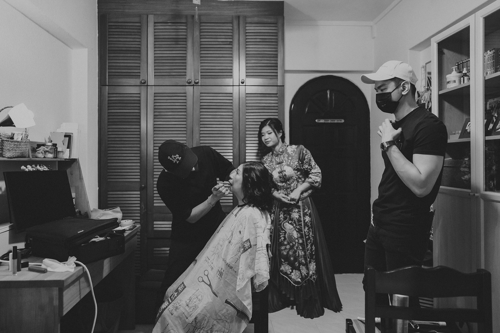 Black and white photo of a barber giving a haircut to a woman in a salon, with a woman in a traditional dress and a man in a baseball cap watching.