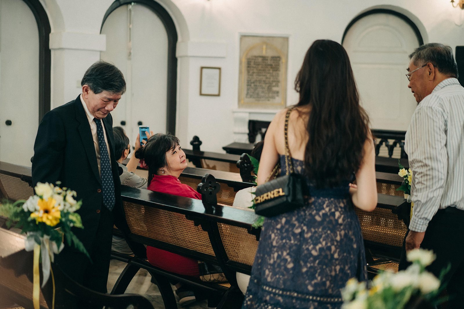A formal gathering in a church, with guests in elegant attire seated in pews. An elder man in a suit speaks with a woman in a red top, while a woman in a navy lace dress with a Chanel purse stands facing away.