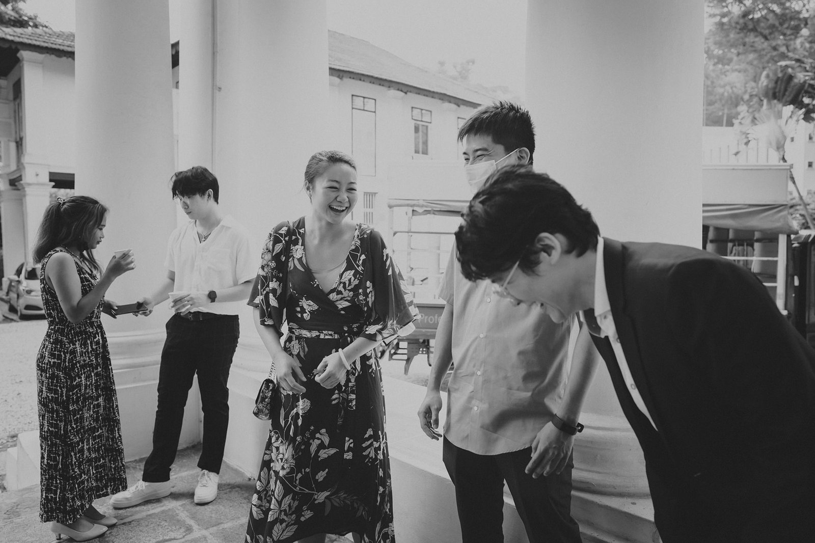 A group of young adults, dressed for a formal occasion, laugh and converse outdoors under a colonnade, with a hint of a street scene in the background.