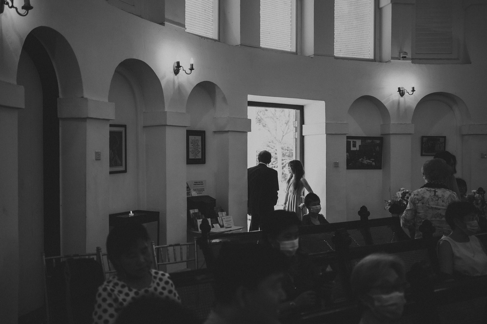 Black and white photo of people in a church, some seated in pews and others standing near an open doorway. A couple stands in the doorway, facing away from the camera.