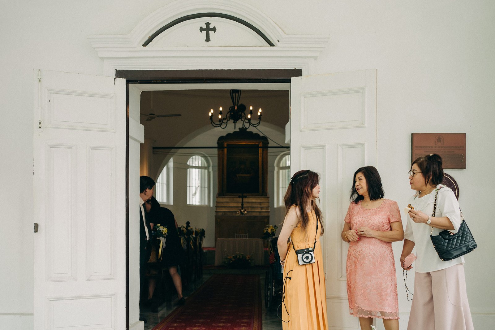 Three women in elegant dresses stand near the open double doors of a white church, with a wedding ceremony visible inside. The woman on the left wears a peach dress, the middle woman a coral lace dress, and the woman on the right a white blouse and pink skirt, holding a black handbag.