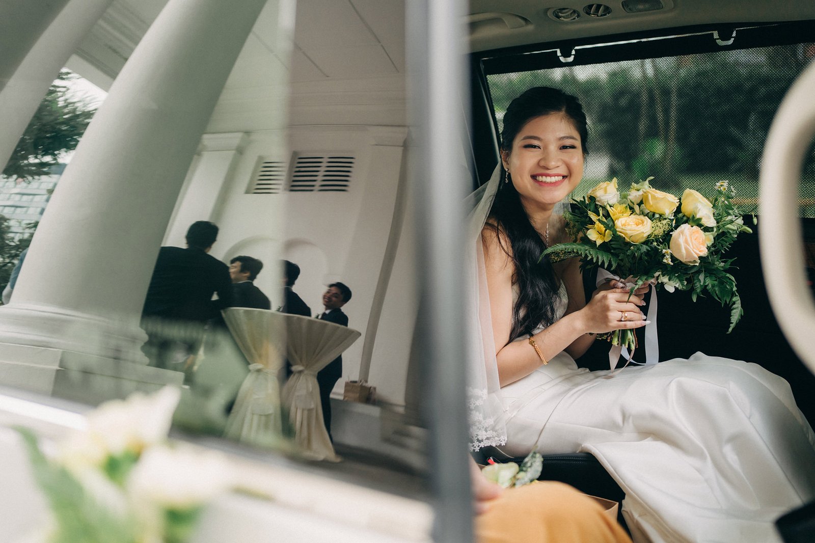 A smiling bride in a white wedding dress and veil sits in the back of a car, holding a bouquet of yellow and white roses. Reflections of guests and a building are visible in the car window.