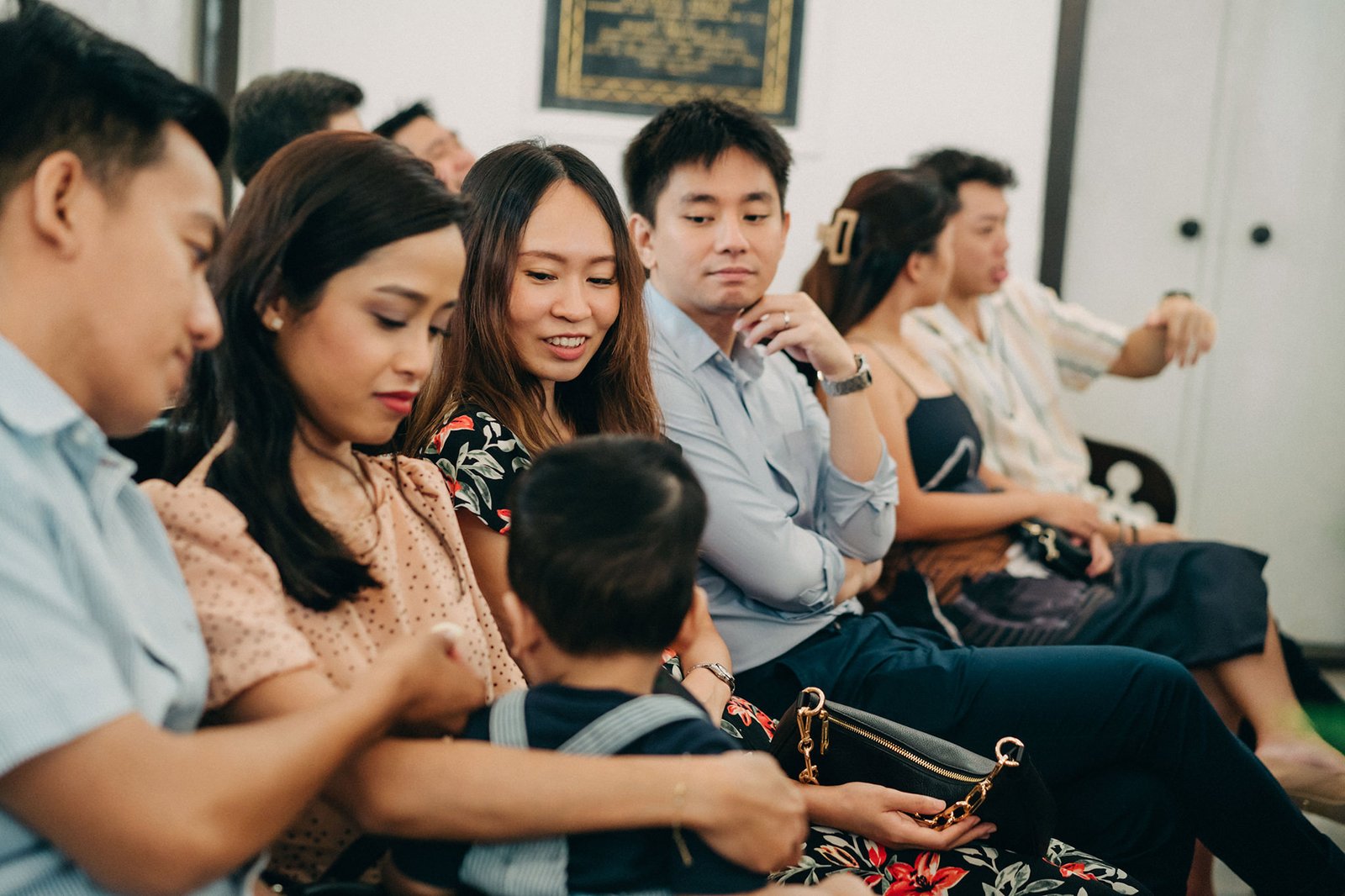 A diverse group of people, including a young child in the foreground, are seated in rows indoors, attentively watching something out of frame. The adults wear smart casual attire, with women in floral prints and men in button-down shirts.