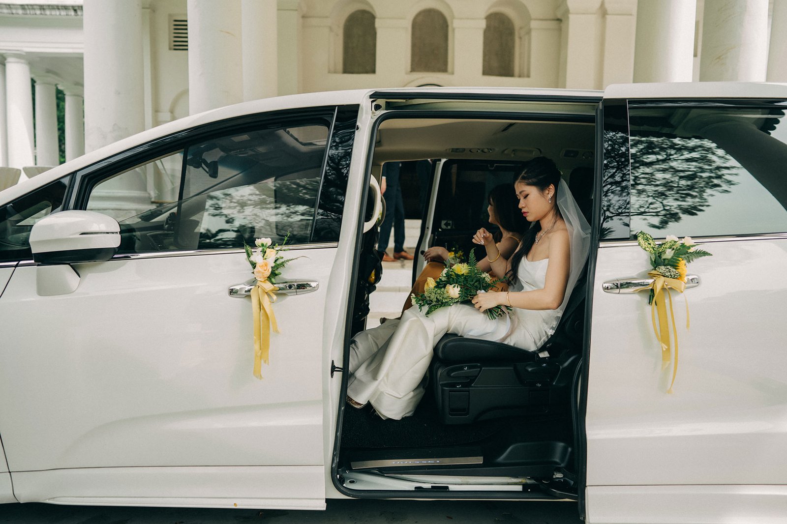 A bride in a white wedding dress and veil sits in the open door of a white minivan, holding a bouquet of white and green flowers. The vehicle is decorated with yellow ribbons and floral accents.