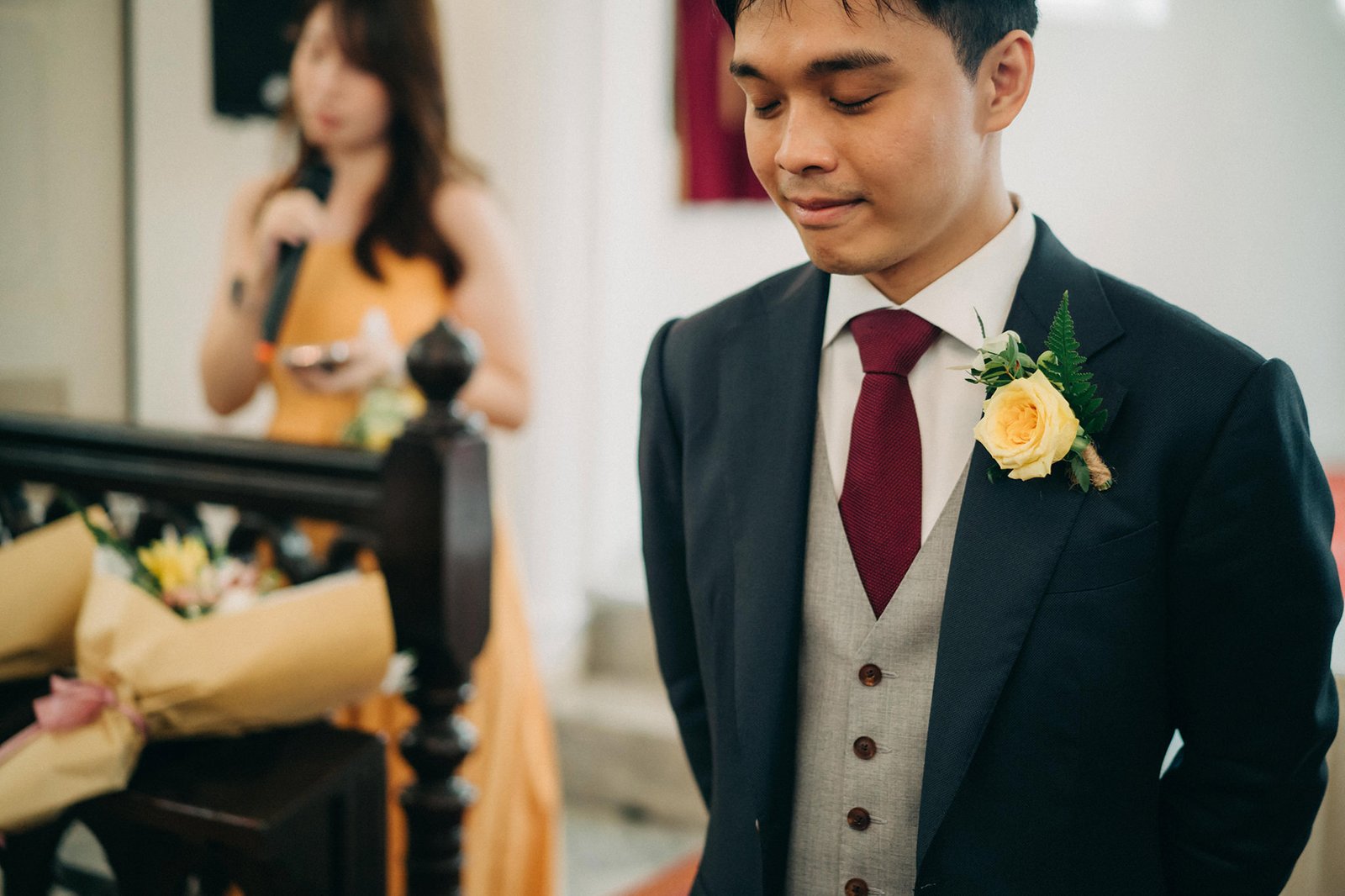 Groom in a dark suit, light grey vest, and burgundy tie with a yellow rose boutonniere stands with eyes closed. A woman in an orange dress holds a microphone in the blurred background.