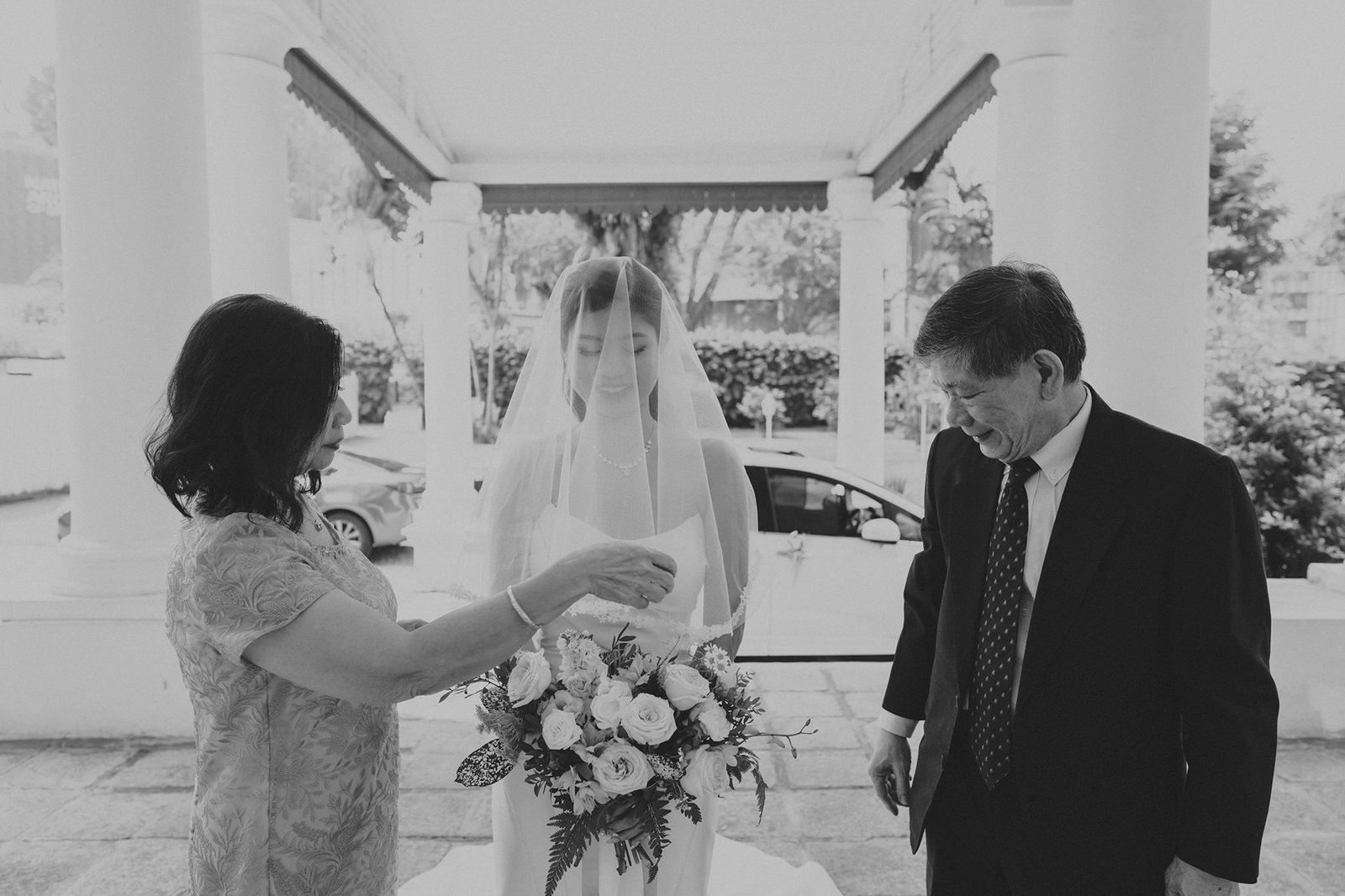 A bride in a veil holds a bouquet while a woman adjusts her dress, and a man in a suit looks on warmly. This scene captures a touching pre-wedding moment.