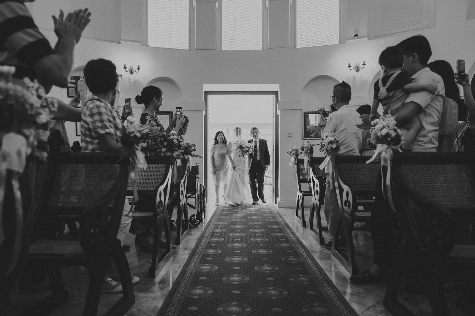 Bride and groom walk down the aisle in a church, guests capture the moment with phones at Armenian Church Singapore. A wedding ceremony in a bright, airy venue.