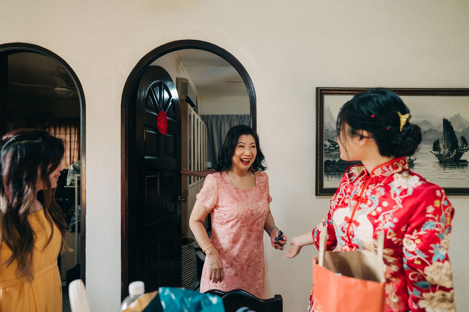 A joyful woman in a pink dress greets guests at a doorway, while another woman in a red traditional Chinese dress smiles and reaches out. A painting of ships hangs on the wall.