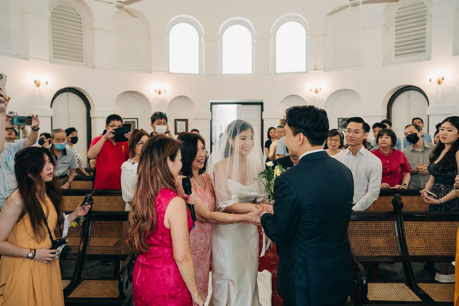 A happy bride in a white wedding dress and veil exchanges vows with her groom in a bright, airy church, surrounded by smiling family and friends.
