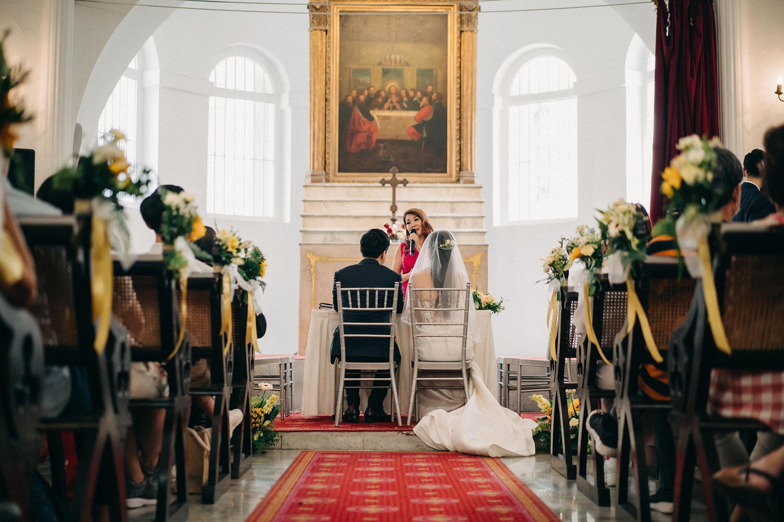 A wedding ceremony in progress, with a bride in a white gown and a groom in a dark suit seated at a table, facing an officiant. Guests are seated in chairs on either side of a red carpet aisle in Armenian Church Singapore.